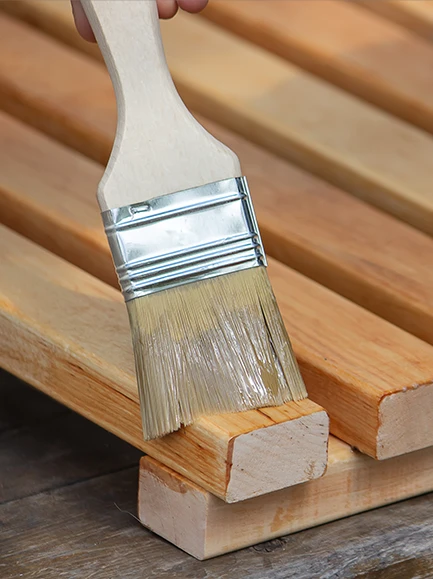 Close-up of a brush applying wood stain to wooden planks for outdoor furniture or decking.