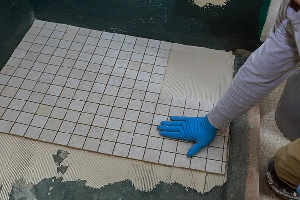 Worker installing small square tiles on a floor using adhesive mortar.