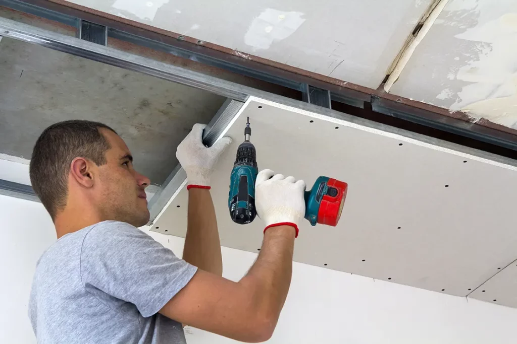 Worker installing drywall on the ceiling with a power drill.