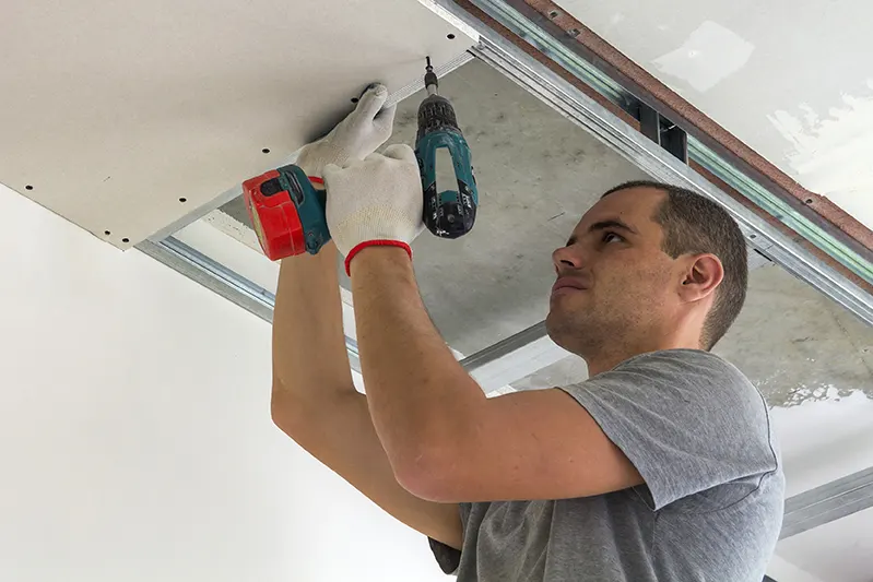 Worker installing drywall on the ceiling with a power drill.