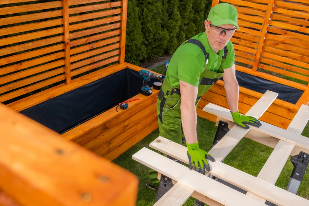 Worker building custom wooden planter boxes with privacy screens in a backyard.