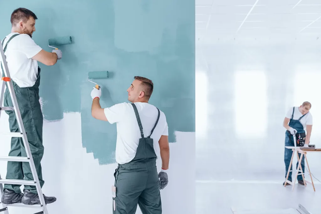 Two workers applying a fresh coat of blue paint on a wall with rollers, while another prepares tools in the background.