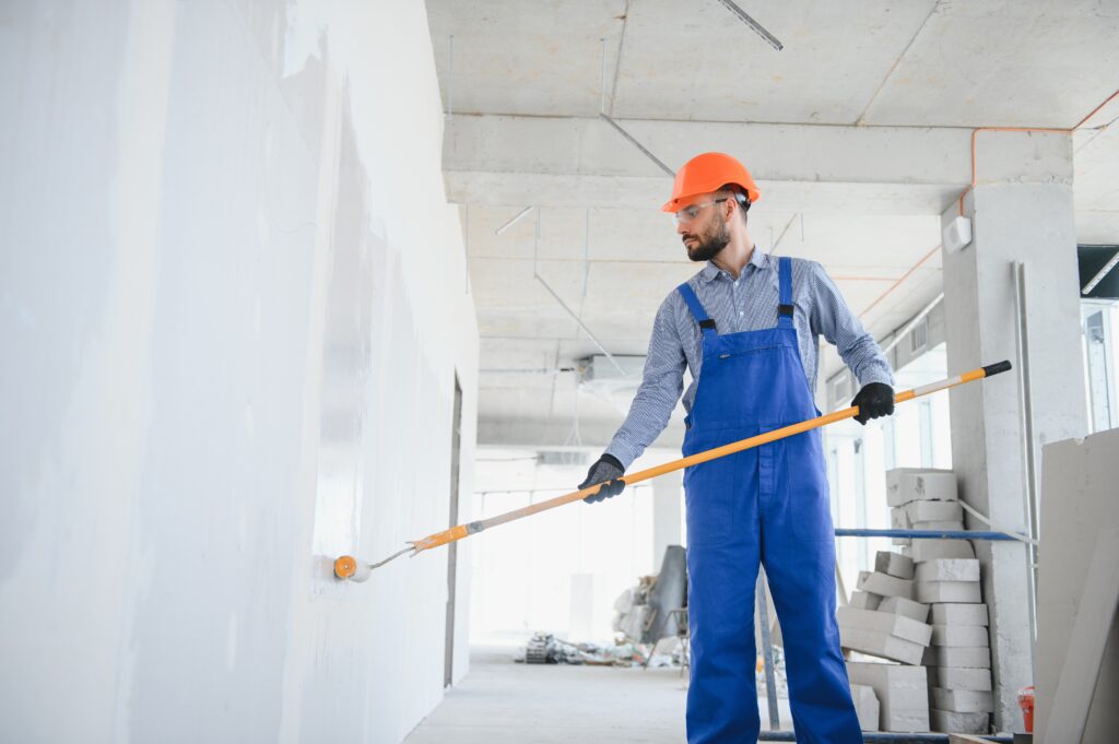 Construction worker in a blue overall applying paint to a wall with a roller.