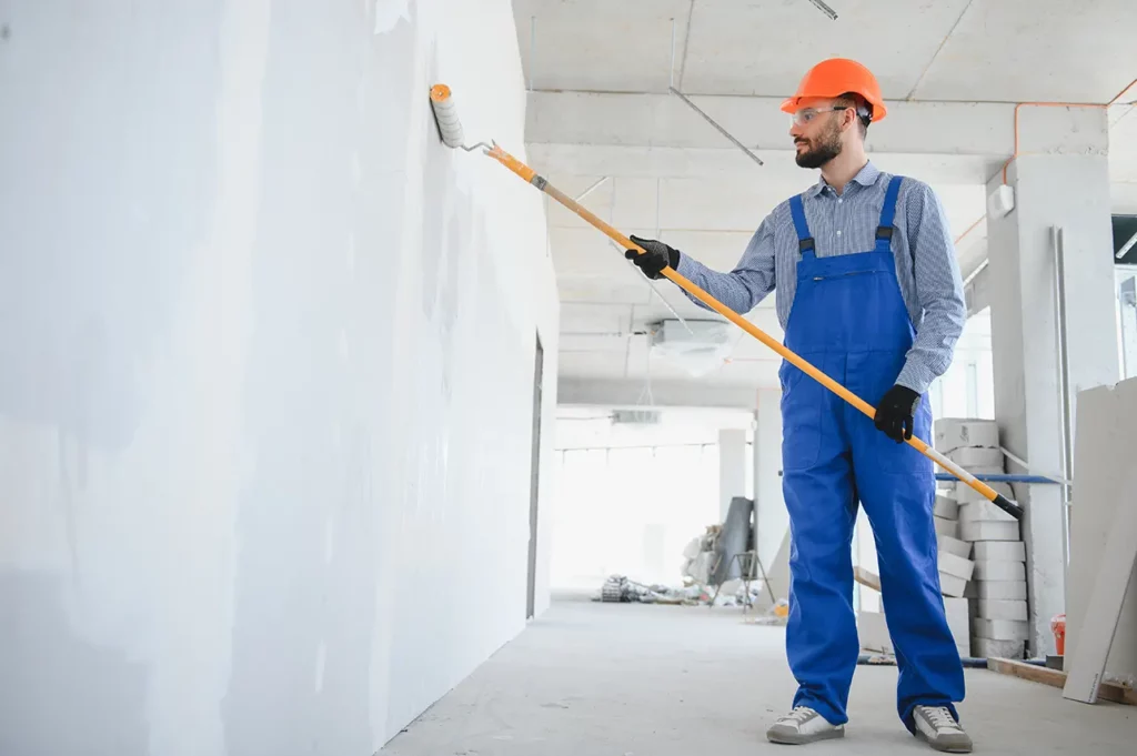 Worker applying white paint to a wall using a roller during an interior painting project.