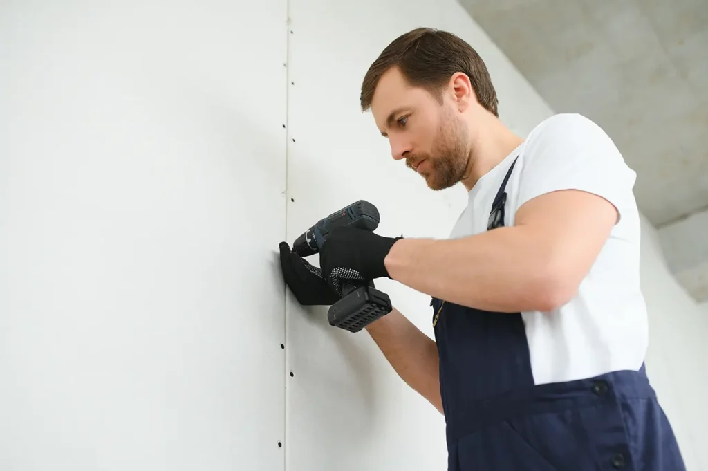 Worker installing drywall with a power drill, focusing on precision.