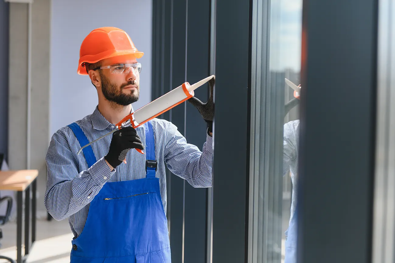 Worker applying sealant to a window frame with a caulking gun during trim installation.