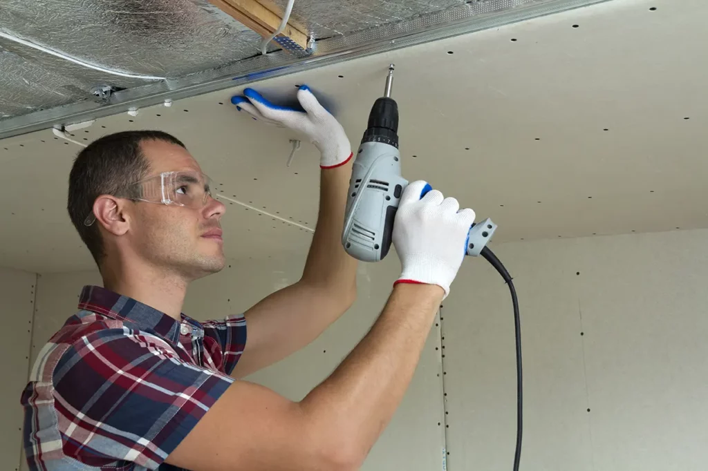 Worker securing drywall to ceiling with a power drill.