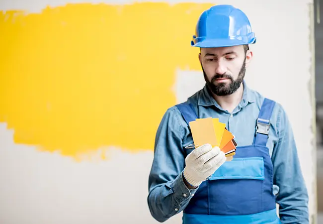 Painter in safety gear holding color swatches, selecting paint for a room with a yellow accent wall, showcasing Coca Construction's attention to detail.