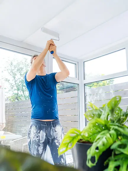 Man painting a ceiling in a sunroom with large windows using a roller.