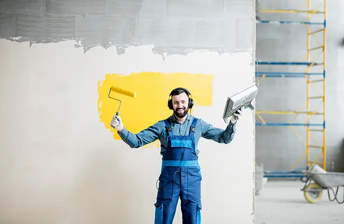 Commercial painter in protective gear holding a roller brush and plastering tool with scaffolding in the background.