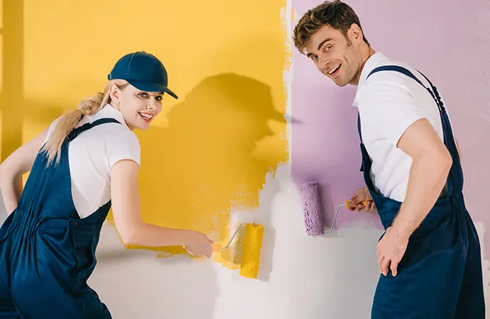 Two painters smiling while painting a wall with yellow and purple colors.