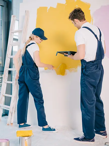 Two professional painters in overalls painting a wall with yellow and purple colors.