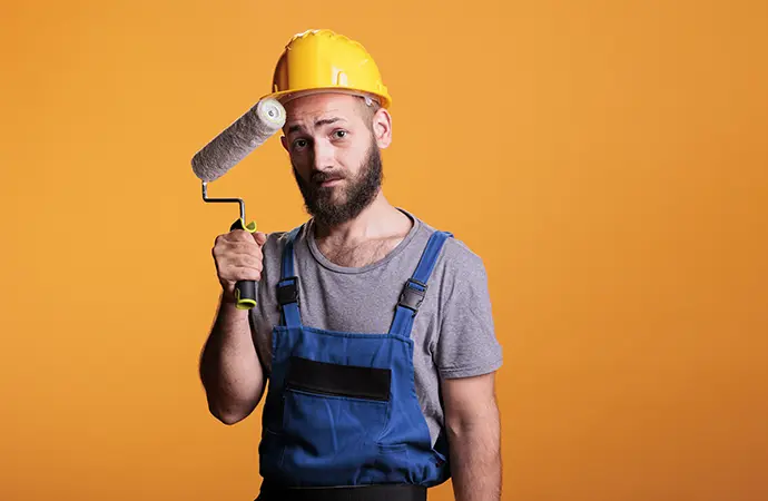 Bearded house painter in a hard hat holding a paint roller against an orange background, representing Coca Construction's professional painting services.