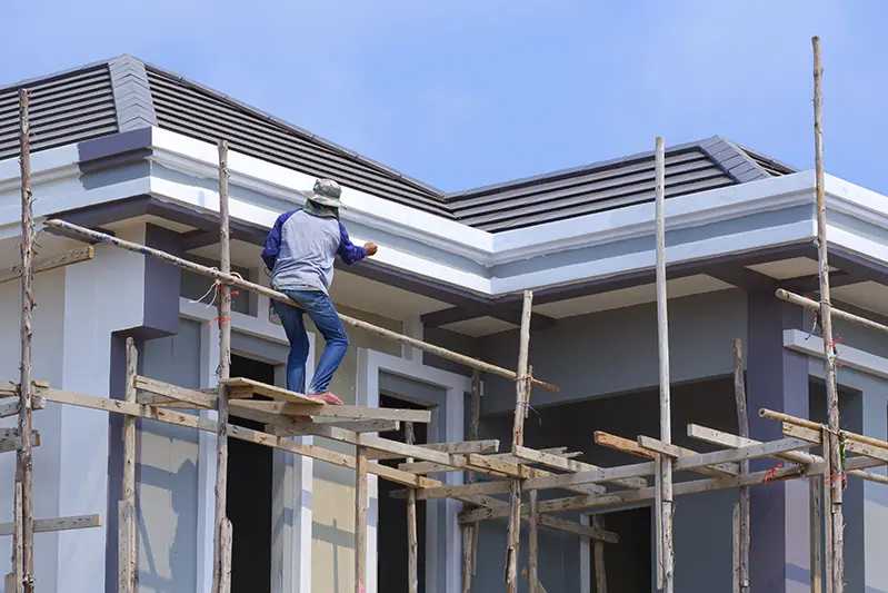 House painter on scaffolding applying fresh paint to the exterior of a modern home under a clear blue sky.