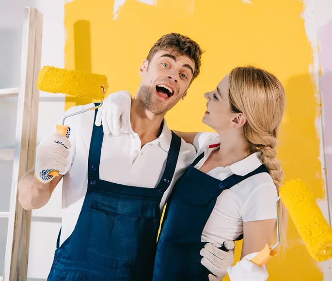 Happy couple painting a room together with yellow paint rollers, wearing blue overalls during a house painting project.