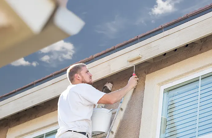 Professional painter on a ladder applying fresh paint to the exterior trim of a house under a clear sky.