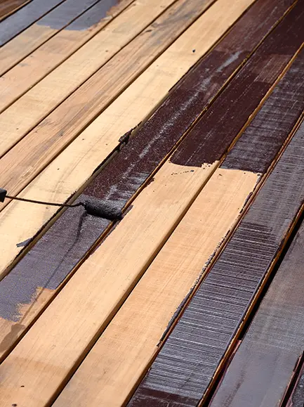 Wooden deck being stained with a dark brown color using a paint roller.