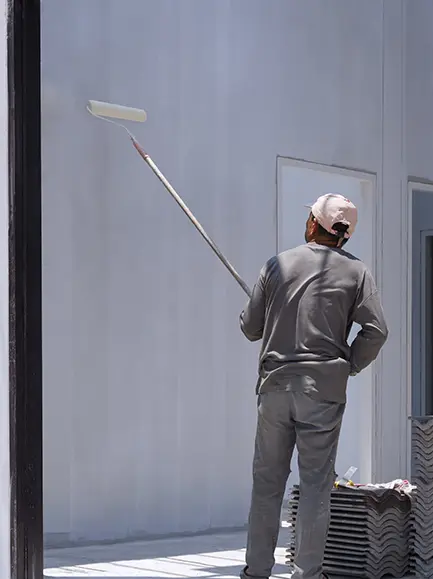 Professional painter applying white paint to an exterior wall using a long-handled roller.