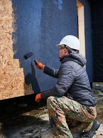 Worker in protective gear painting a building’s exterior wall with a roller.