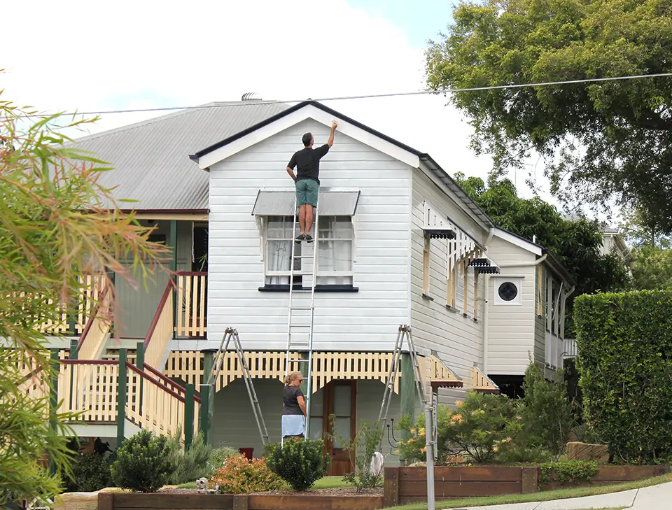 Two men painting the exterior of a two-story house using a ladder, focusing on the top facade.